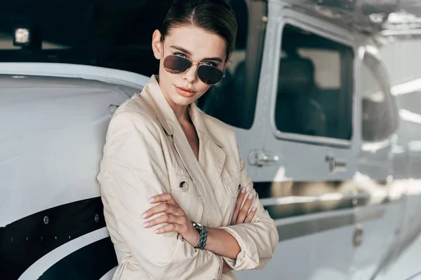 Jeune femme en lunettes de soleil et veste posant avec les bras croisés et regardant la caméra près de l'avion — Photo de stock