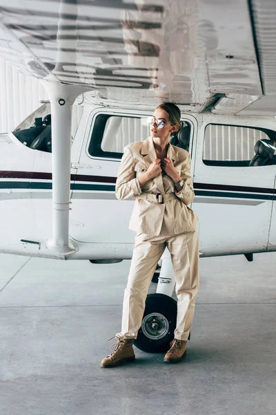 Attraente donna elegante in occhiali da sole e giacca posa vicino aereo in hangar — Foto stock