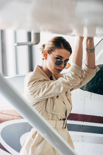 Jeune femme en lunettes de soleil et veste regardant la caméra près de l'avion — Photo de stock