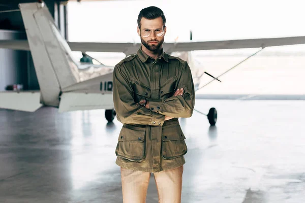 Confident fashionable young man in green jacket and eyeglasses posing with crossed arms near airplane in hangar — Stock Photo