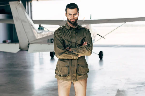 Confiante jovem na moda em casaco verde e óculos posando com braços cruzados perto de avião no hangar — Fotografia de Stock