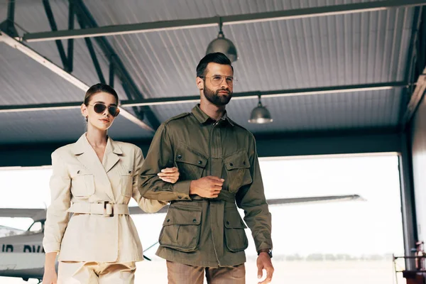 Низкий угол зрения на модную молодую пару в куртках, идущую рядом с самолетом в ангаре — стоковое фото