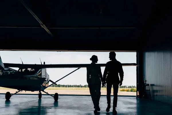 Visão traseira de silhuetas de jovem casal andando perto de avião em hangar — Fotografia de Stock