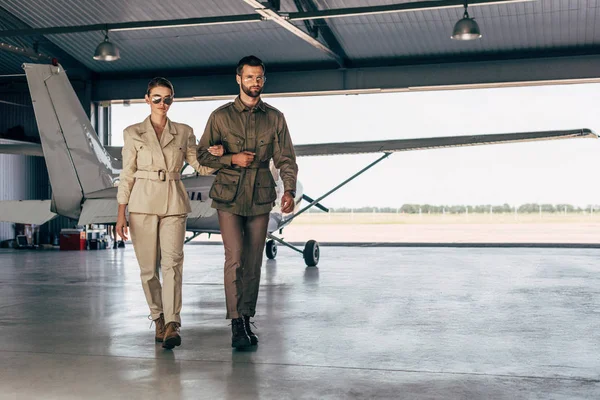 Confiante jovem casal na moda em jaquetas andando perto de avião no hangar — Fotografia de Stock