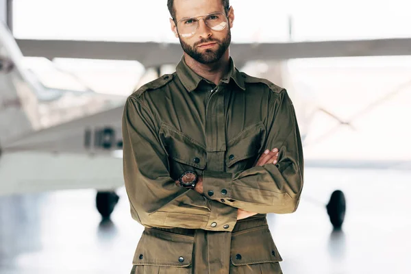 Joven guapo en chaqueta verde y anteojos de pie con los brazos cruzados cerca del avión - foto de stock