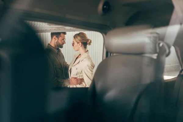 Enfoque selectivo de la joven pareja de moda en chaqueta abrazándose y mirándose el uno al otro cerca del avión - foto de stock