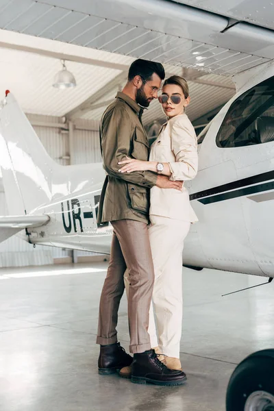Stylish young couple in jackets embracing near airplane in hangar — Stock Photo