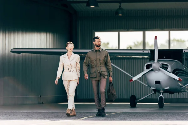 Bell'uomo che trasporta borsa e cammina con elegante ragazza vicino hangar con aereo — Foto stock