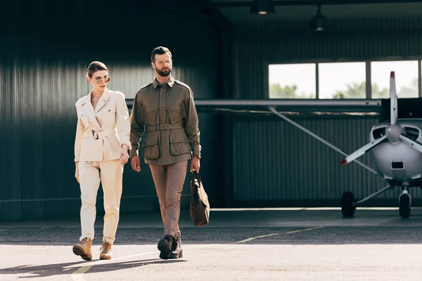 Uomo serio che trasporta borsa e cammina con ragazza alla moda vicino hangar con aereo — Foto stock