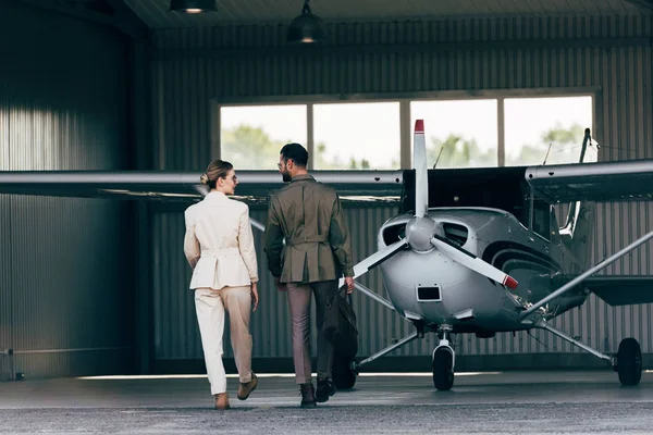 Vue arrière de l'homme portant sac et marche avec petite amie élégante près du hangar avec avion — Photo de stock