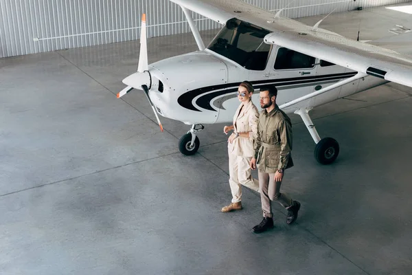 Blick aus der Vogelperspektive auf ein modisches junges Paar in stylischen Jacken, das im Hangar mit Flugzeug spaziert — Stockfoto