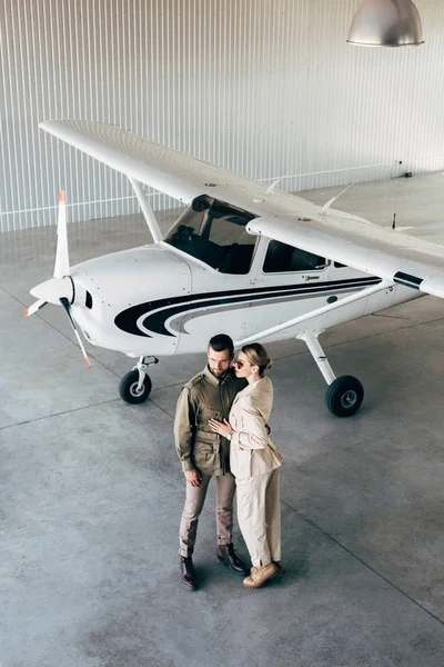 Visão de alto ângulo de casal jovem na moda em jaquetas elegantes abraçando no hangar com avião — Fotografia de Stock