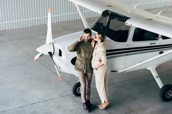 Blick aus der Vogelperspektive auf ein modisches junges Paar in schicken Jacken, das in der Nähe eines Flugzeugs im Hangar steht — Stockfoto