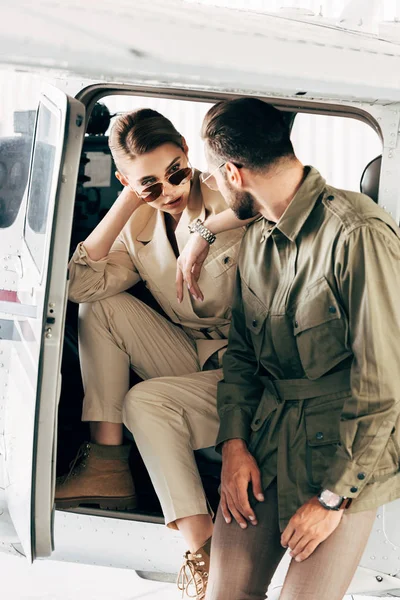 Pareja joven de moda en chaquetas mirándose cerca del avión - foto de stock