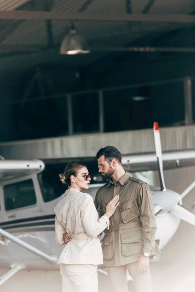 Fashionable young couple in stylish jackets standing in hangar with airplane — Stock Photo