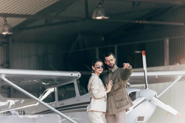 Schöner Mann in Jacke zeigt mit dem Finger auf stylische Freundin in Flugzeugnähe im Hangar — Stockfoto