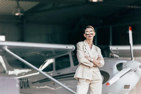 Стильна молода жінка в сонцезахисних окулярах і піджаках позує схрещеними руками біля літака — стокове фото