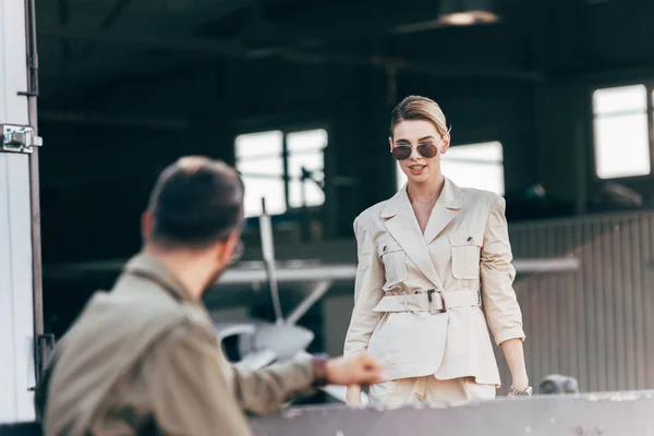 Messa a fuoco selettiva di bella giovane donna in occhiali da sole e giacca a piedi al fidanzato vicino hangar con aereo — Foto stock