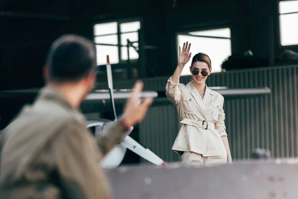 Selective focus of stylish woman in sunglasses waving by hand to boyfriend near plane — Stock Photo