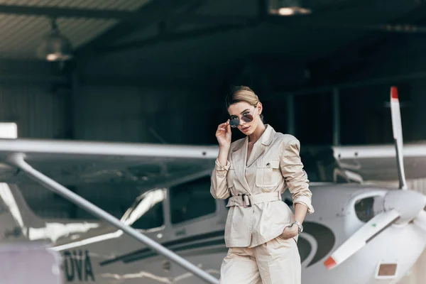 Attractive young woman in sunglasses and jacket posing near aircraft — Stock Photo