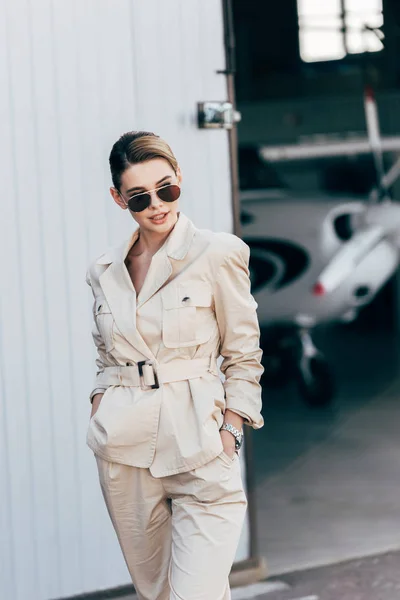 Молода красива жінка в сонцезахисних окулярах і піджак позує руками в кишенях біля літака — стокове фото