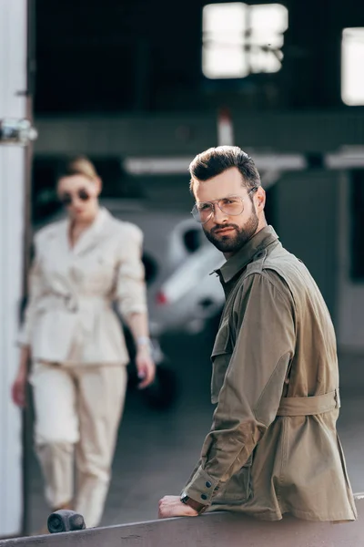 Enfoque selectivo de hombre con estilo en gafas y chaqueta mirando a la cámara mientras su novia caminando cerca del hangar con avión - foto de stock