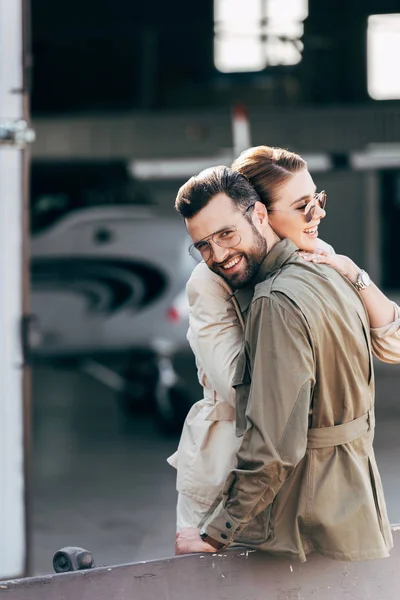 Hombre feliz en gafas y chaqueta mirando a la cámara mientras abraza novia cerca del hangar con avión - foto de stock