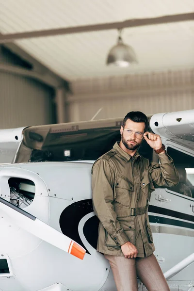 Jovem bonito em casaco verde e óculos olhando para longe perto do avião — Fotografia de Stock