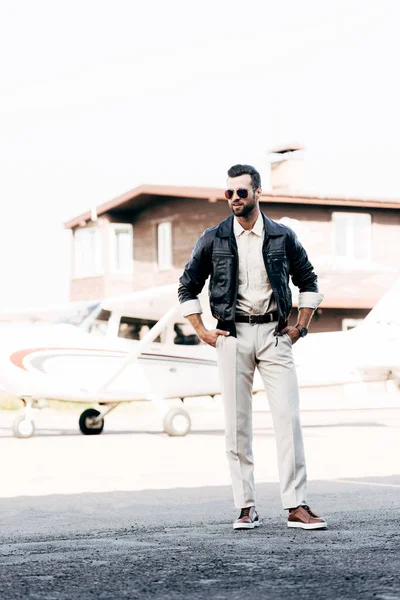 Sonriente piloto masculino de moda en chaqueta de cuero y gafas de sol de pie con las manos en la cintura cerca del avión - foto de stock