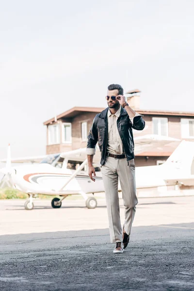 Hombre elegante guapo en chaqueta de cuero y gafas de sol caminando cerca de avión - foto de stock