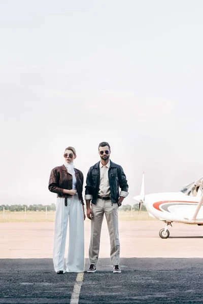 Jovem casal na moda em óculos de sol e jaquetas de couro de pé perto do avião — Fotografia de Stock