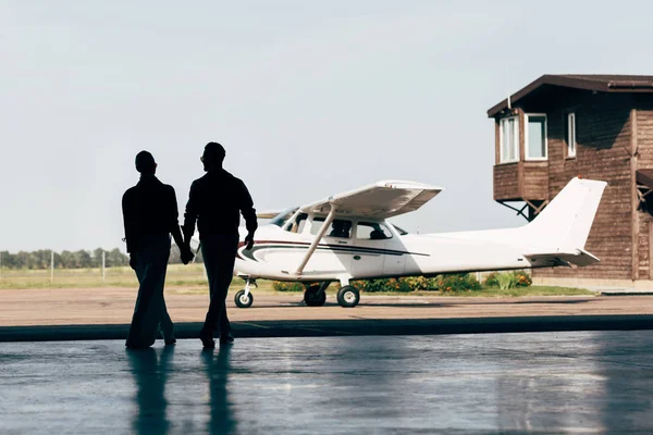 Vista posteriore di sagome di coppia elegante che si tiene per mano e cammina nell'hangar vicino all'aereo — Foto stock