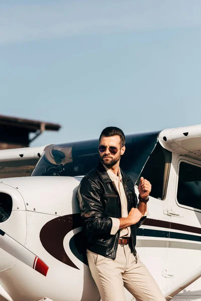 Pilota maschio in giacca di pelle e occhiali da sole guardando lontano vicino all'aereo — Foto stock