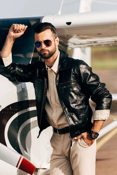 Piloto masculino serio de moda en chaqueta de cuero y gafas de sol de pie cerca del avión — Stock Photo