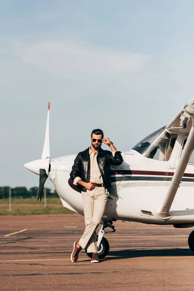 Piloto masculino con estilo en chaqueta de cuero y gafas de sol posando cerca de aviones — Stock Photo