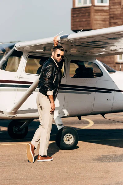 Guapo piloto masculino con estilo en chaqueta de cuero y gafas de sol de pie cerca de los aviones - foto de stock