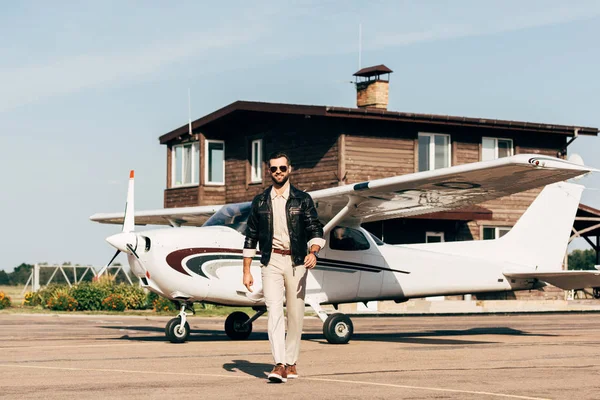 Jovem piloto masculino na moda em jaqueta de couro e óculos de sol andando perto do avião — Fotografia de Stock