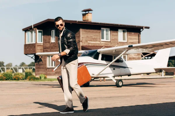 Стильный молодой человек в кожаной куртке и солнцезащитных очках ходьба с чемоданом рядом с самолетом — стоковое фото