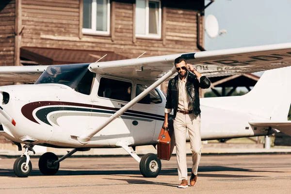 Модний чоловік у шкіряній куртці та сонцезахисних окулярах, що ходять з валізою біля літака — стокове фото