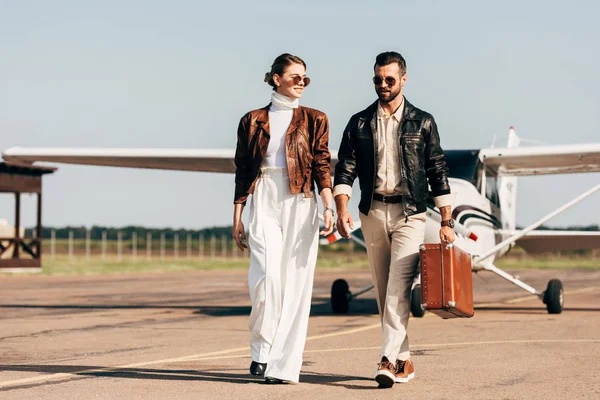 Happy young couple in leather jackets and sunglasses walking with retro suitcase near airplane — Stock Photo