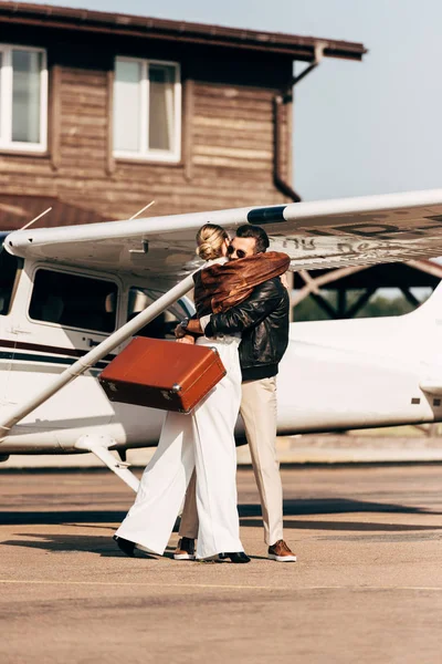 Junges Paar in Lederjacken mit Vintage-Koffer umarmt sich in Flugzeugnähe — Stockfoto