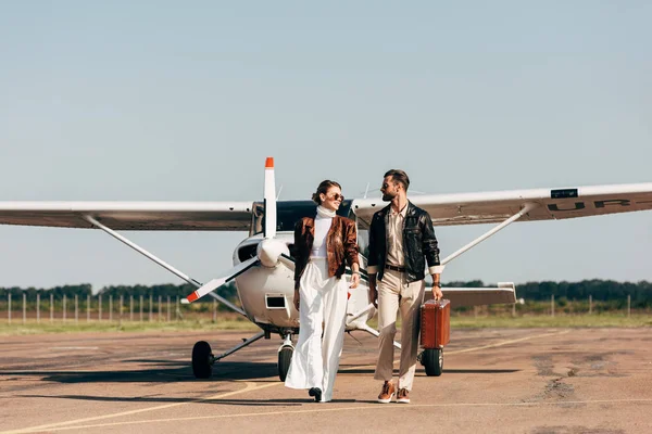 Elegante pareja joven en chaquetas de cuero y gafas de sol caminando con maleta retro cerca del avión - foto de stock