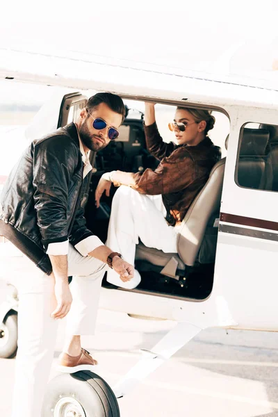 Jeune homme en lunettes de soleil et veste en cuir regardant la caméra tandis que sa petite amie assise dans l'avion — Photo de stock