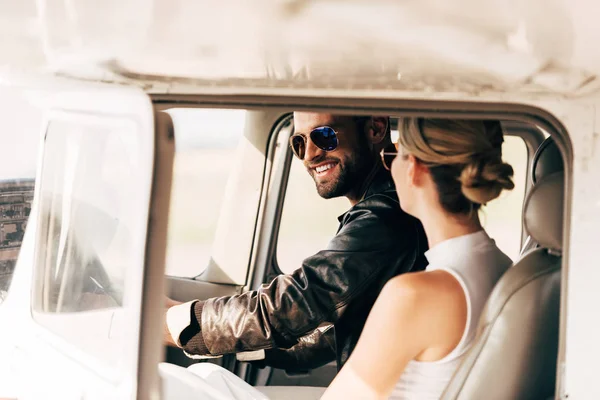 Pilota maschio sorridente in occhiali da sole e giacca di pelle con la sua ragazza seduta vicino in cabina di aeroplano — Foto stock