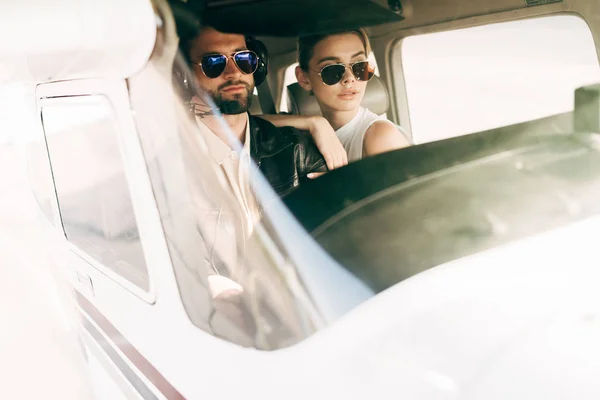 Piloto masculino serio en auriculares y gafas de sol sentado con novia en cabina de avión - foto de stock