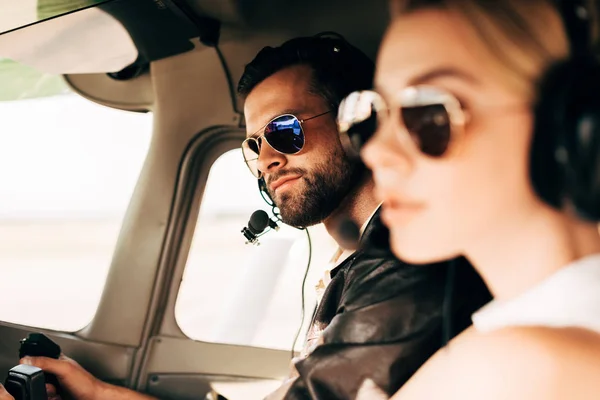 Enfoque selectivo de piloto masculino guapo en auriculares y gafas de sol sentado con novia en cabina de avión - foto de stock