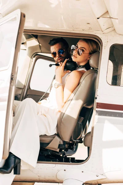 Atractiva mujer joven en gafas de sol y auriculares sobre el cuello sentado cerca de novio en cabina de avión - foto de stock