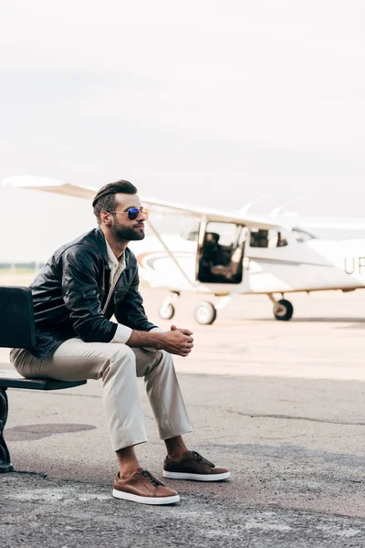 Elegante joven piloto masculino en gafas de sol sentado en el banco cerca de avión - foto de stock