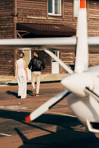 Vista trasera de mujer joven caminando con novio en chaqueta de cuero cerca de avión - foto de stock