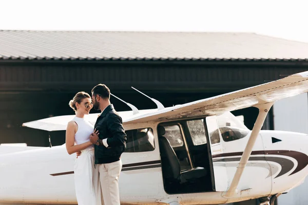 Stylish couple in sunglasses embracing and standing face to face near airplane — Stock Photo